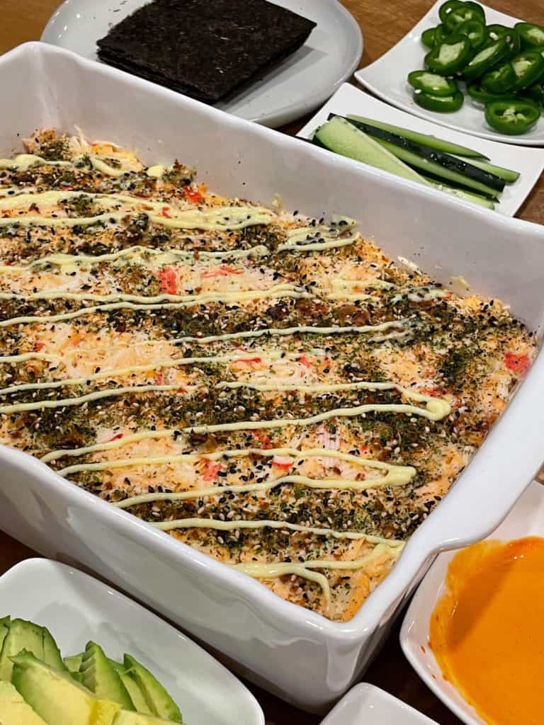 Salmon sushi bake in a white baking dish drizzled with creamy mayo and furikake, with nori and cucumbers on the side