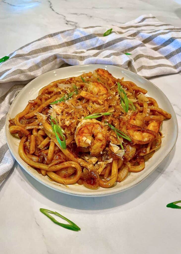 Stir Fried Udon Noodles on a white plate with shrimp garnished with bonito flakes and green onions.
