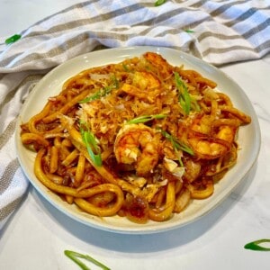 Stir Fried Udon Noodles on a white plate with shrimp garnished with bonito flakes and green onions.