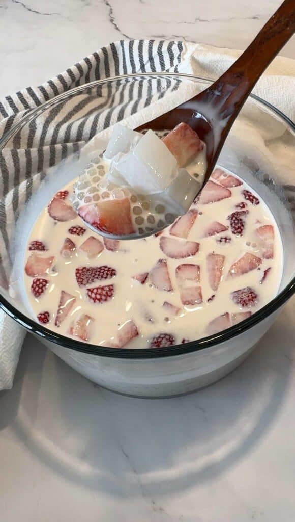Wooden spoon lifting strawberry sago with sago pearls and coconut jelly from a glass bowl of creamy milk and strawberries