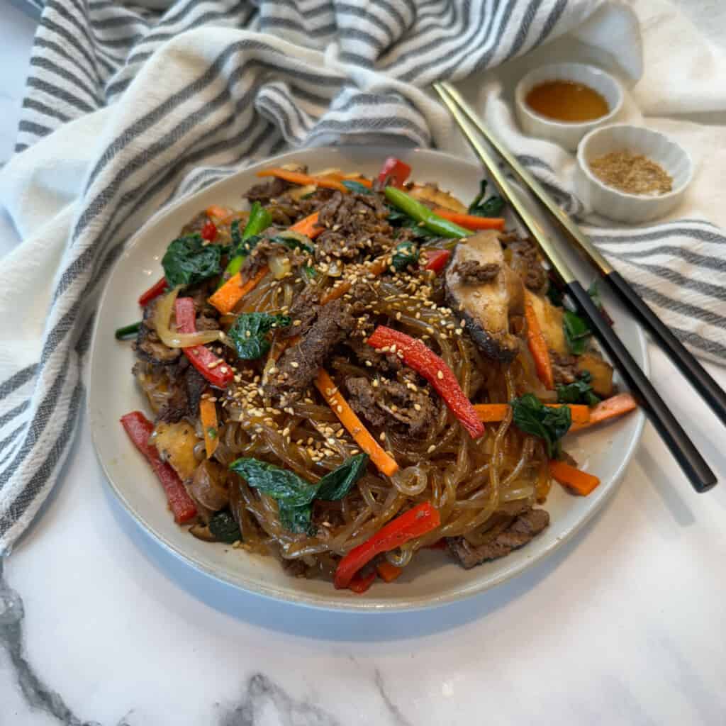 Plate of Korean beef japchae with glass noodles, bulgogi beef, spinach, carrots and peppers topped with sesame seeds