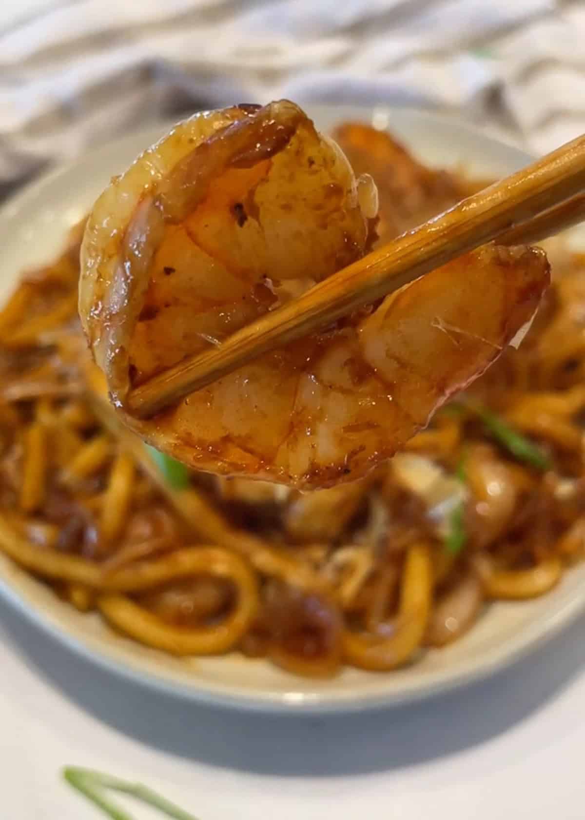 A brown chopstick holding up a cooked shrimp over a plate of yaki udon