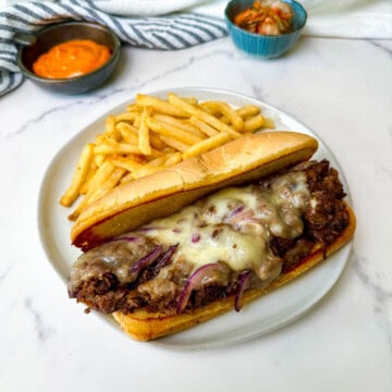 Plated Bulgogi Beef Cheesesteak on a white plate with fries served with condiments.