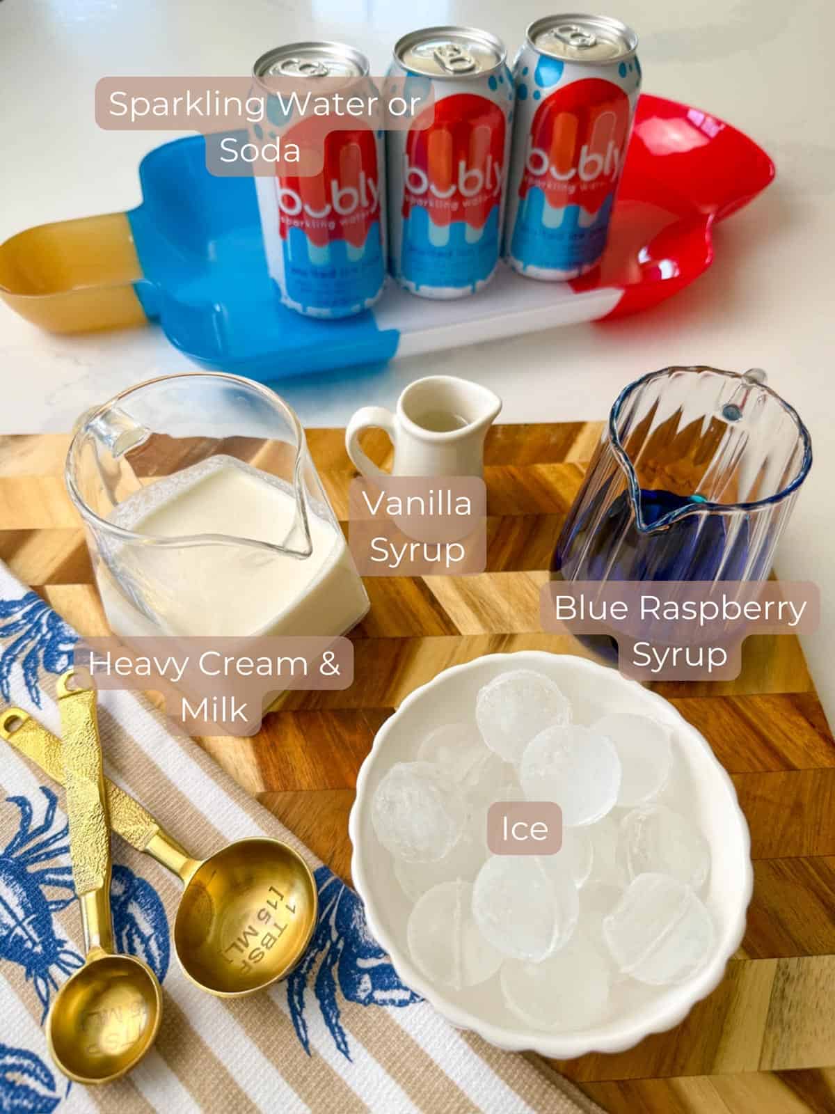 Dirty soda ingredients ice cinnamon bowl milk in a clear glass blue raspberry syrup and a clear glass with the decorative spoon and towel with blue lobsters and beige stripes