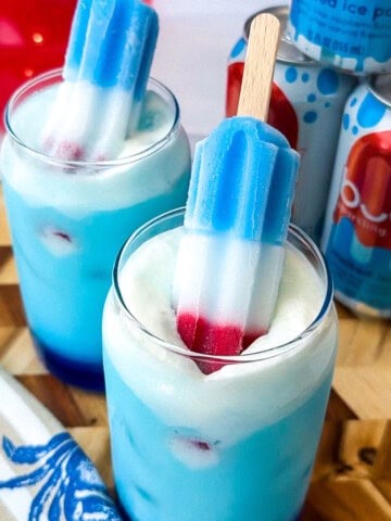 Blue drink and a glass cup with white foam and two rocket shaped popsicles that are red white and blue