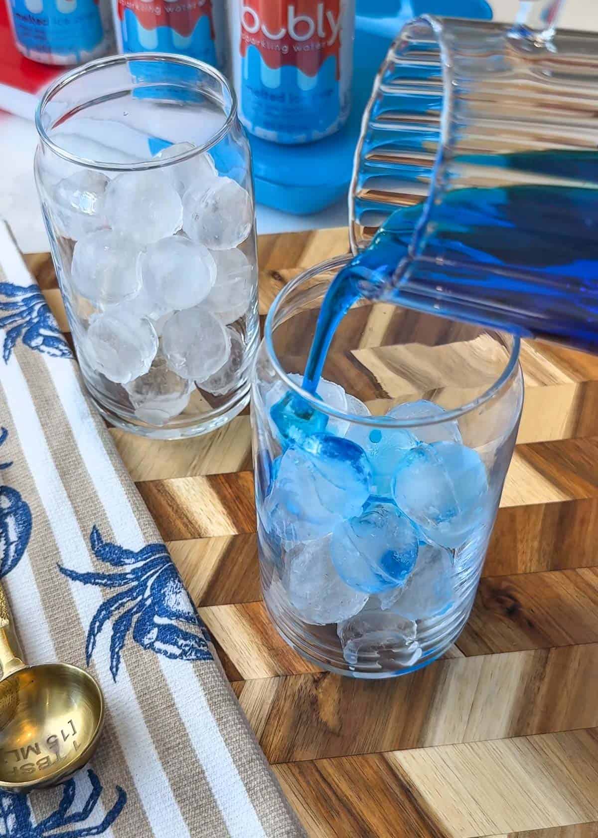 Ice in a clear cup with blue liquid being poured in it with a summer themed background