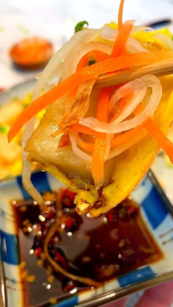 Chopsticks holding fried rice cake with pickled carrots and daikon over soy dipping sauce