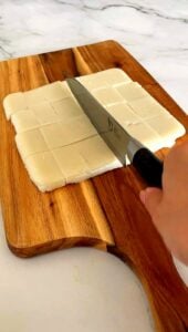 Slicing the chilled steamed rice cake into small rectangles on a wooden cutting board