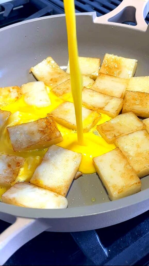 Beaten eggs being poured over golden fried rice cakes in a skillet