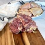 Stack of deboned skin-on chicken thighs on a cutting board with bone-in thighs on a plate behind