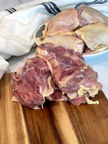 Stack of deboned skin-on chicken thighs on a cutting board with bone-in thighs on a plate behind