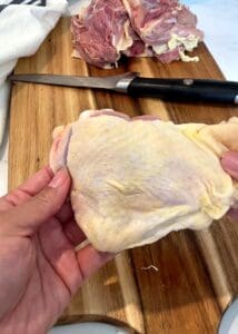 Hand holding a raw skin-on bone-in chicken thigh over a wooden cutting board