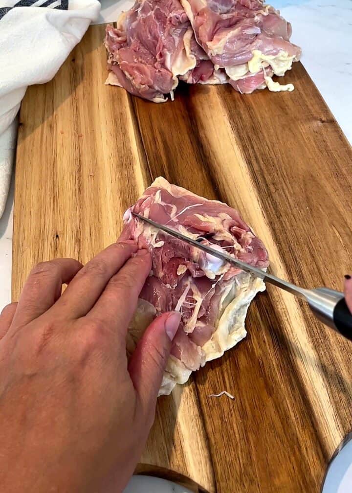 Knife slicing lengthwise along the bone of a raw chicken thigh on a wooden board