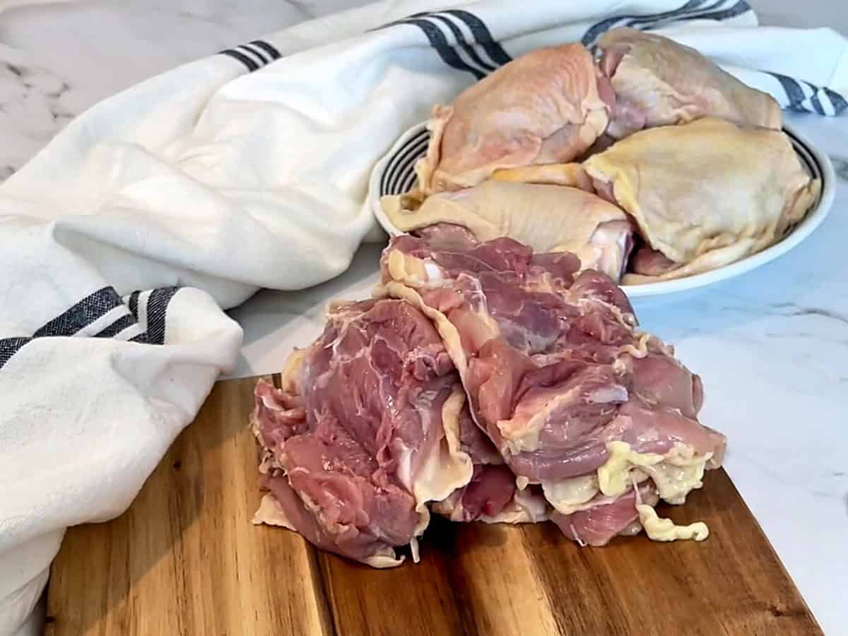 Deboned chicken thighs on a wooden cutting board with a plate of skin-on bone-in thighs behind
