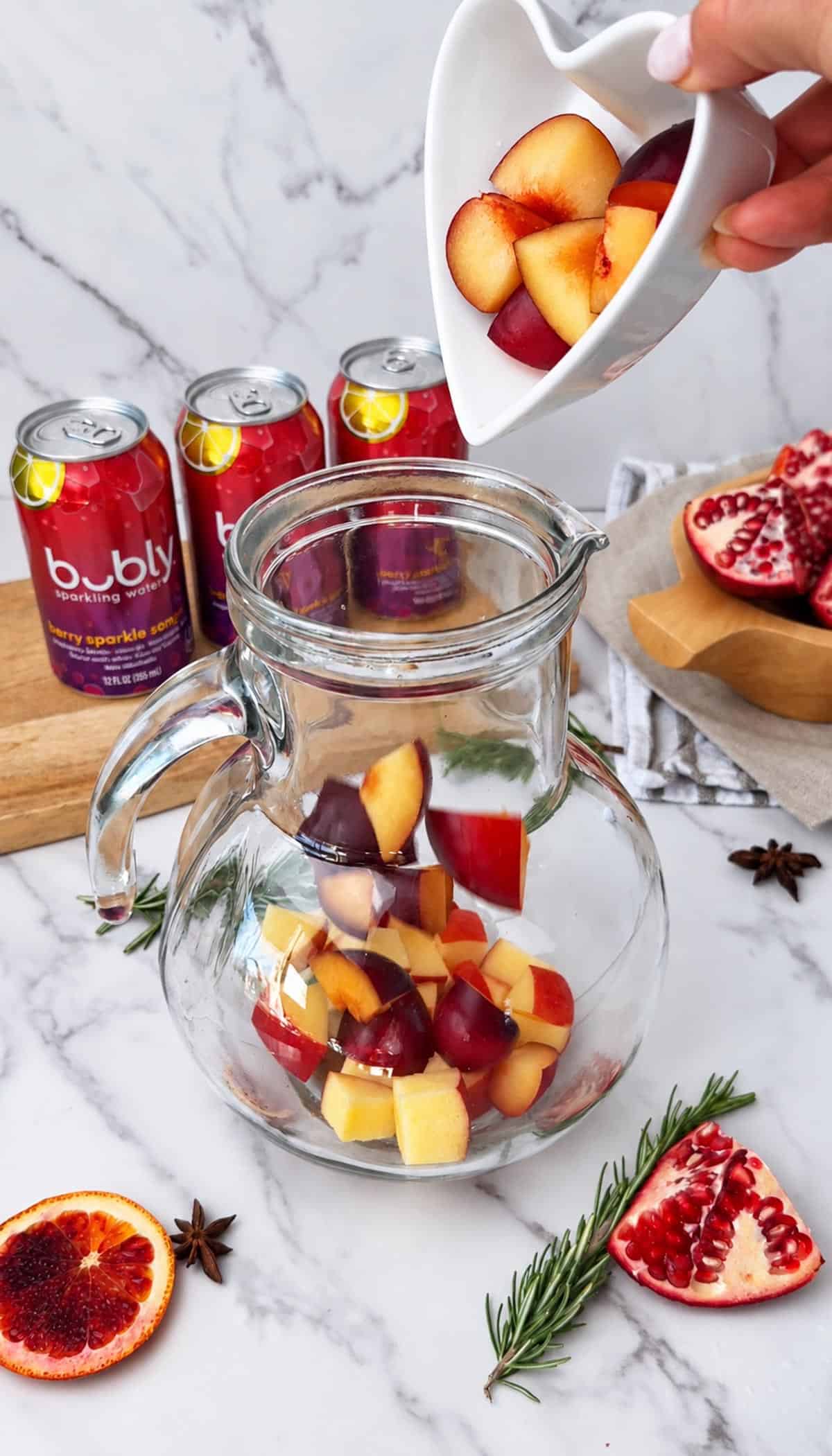 Labeled ingredients for a sangria mocktail, including cranberry pomegranate juice, sparkling water, apples, blood orange, purple plum, pomegranate, rosemary, cinnamon, and star anise.