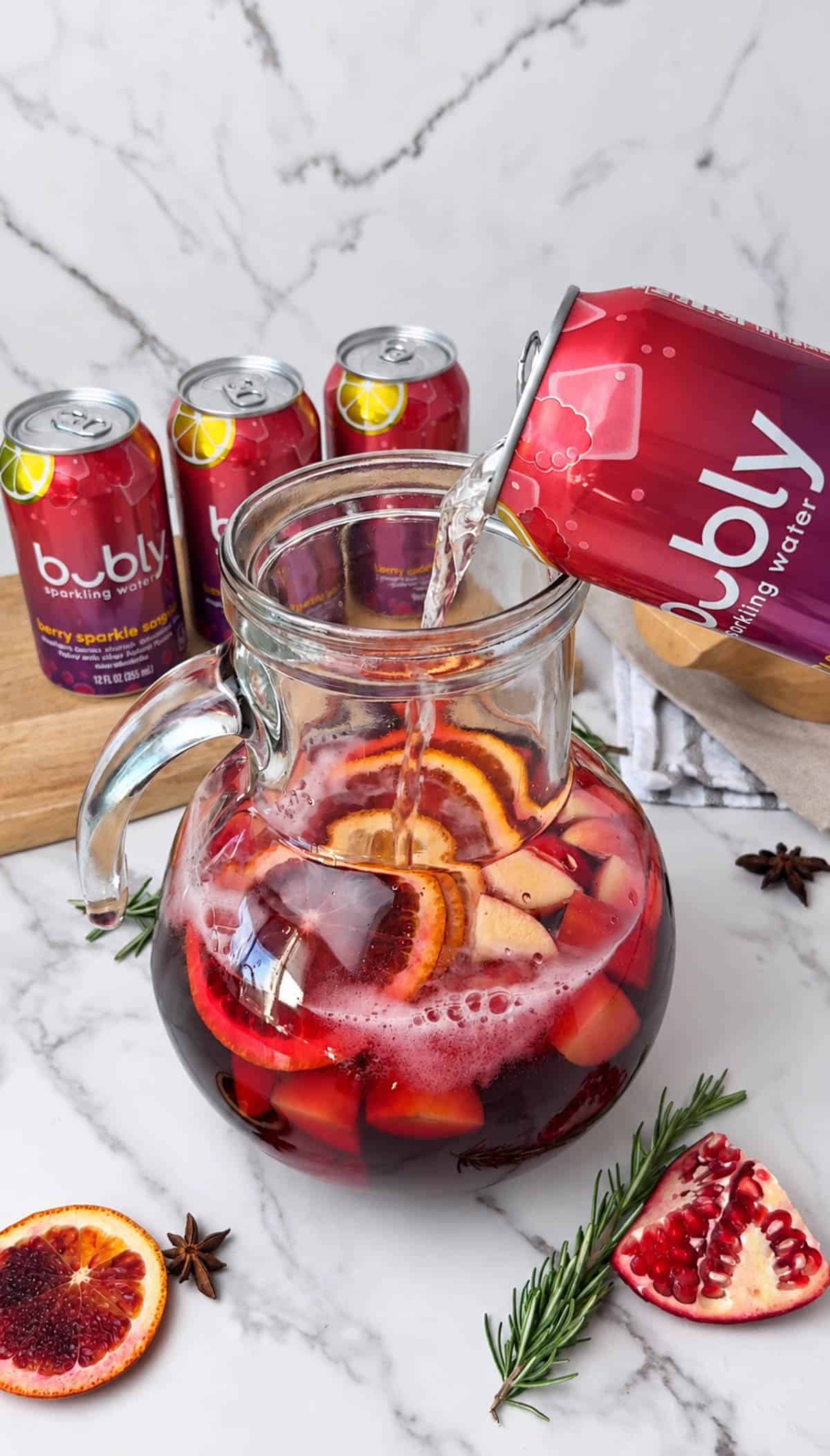 Sparkling water being poured into a pitcher of red sangria mocktail with sliced fruit.