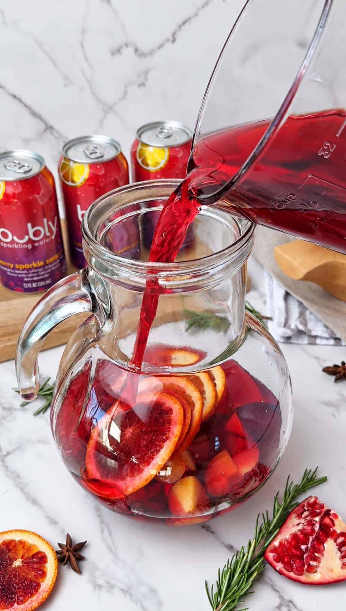 Cranberry pomegranate juice being poured into a pitcher of sliced fruit for a sangria mocktail.