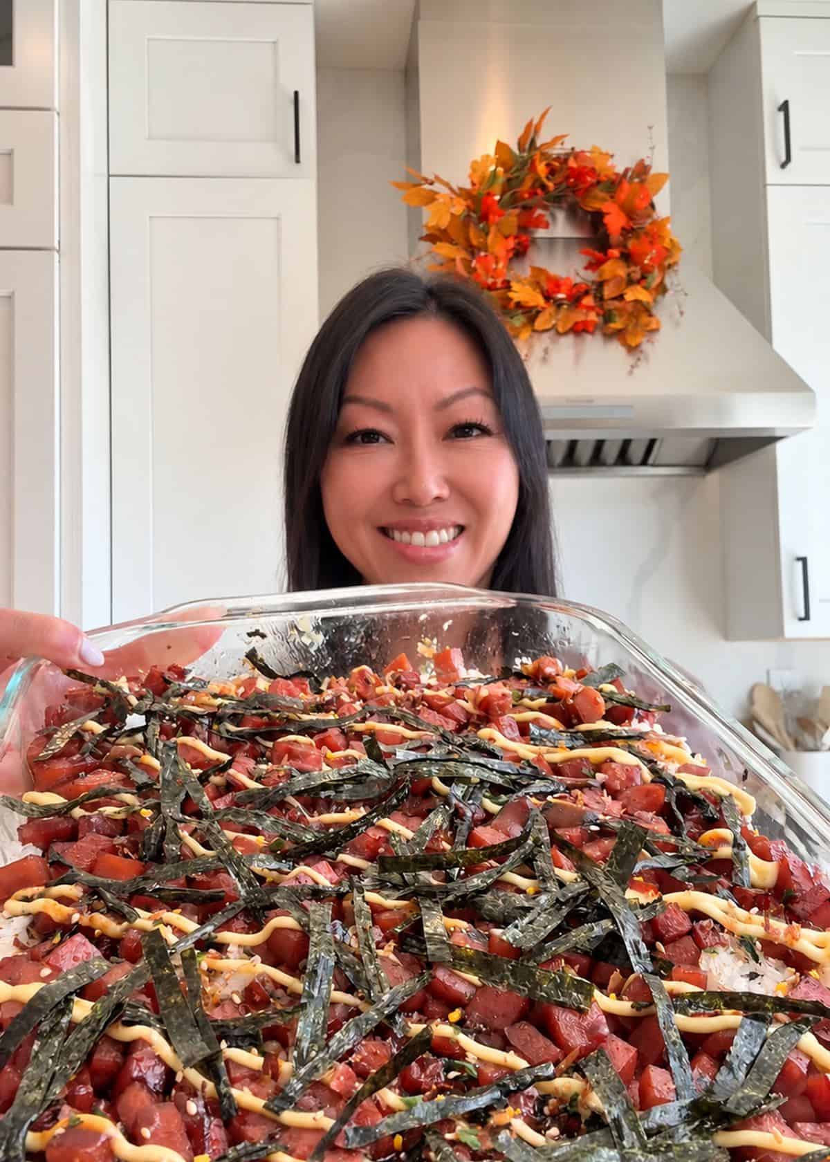 Smiling woman holding a glass baking dish of Spam musubi bake topped with mayo and nori in a kitchen.