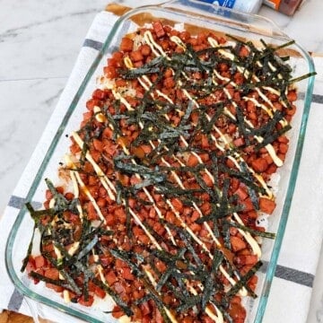 Angled overhead view of a rectangular glass dish filled with Spam musubi bake, drizzled mayo, and shredded nori on white marble.