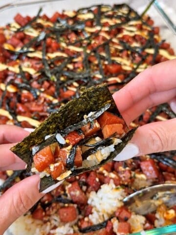 Hands holding a folded piece of nori filled with rice and glazed Spam above a dish of Spam musubi bake.