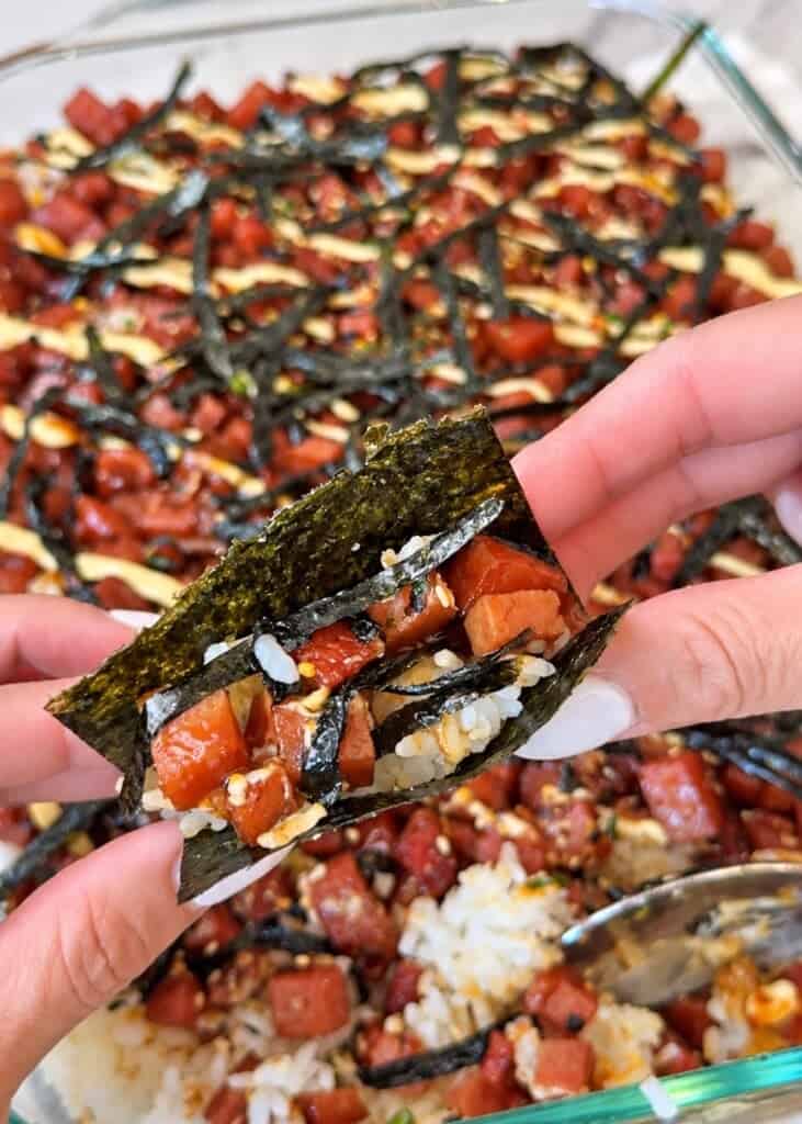 Hands holding a folded piece of nori filled with rice and glazed Spam above a dish of Spam musubi bake.