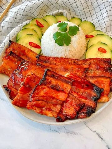 Plate of sticky grilled pork belly slices with white rice, cucumber slices and red chili